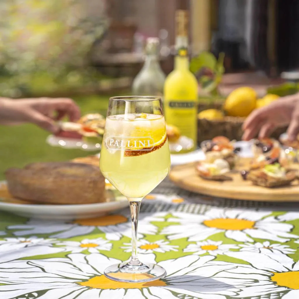 Glass of 0% limoncello with 'Pallini' branding on a floral tablecloth, outdoor setting.